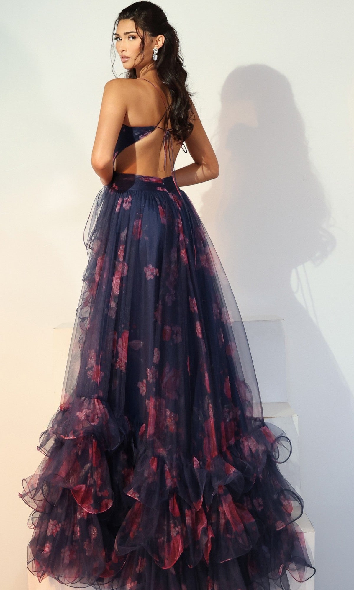 Wearing the Jovani 45712 long prom dress in navy blue and pink floral with ruffles, a woman stands on white steps facing backward, her long wavy hair flowing and statement earrings completing the elegant look.